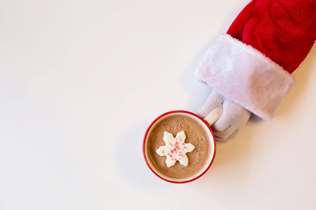 A festive hot cocoa topped with a snowflake marshmallow, held by a Santa gloved hand on a white background.