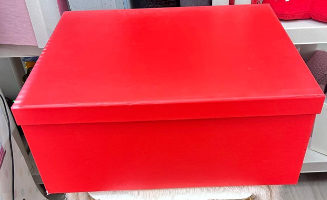 Caja roja grande con tapa cerrada sobre una superficie, fondo con estantes desenfocados.